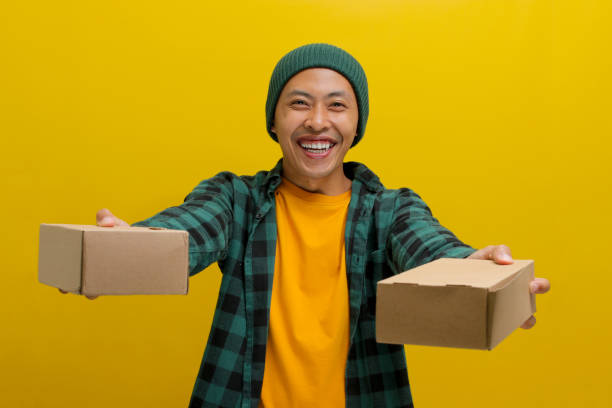 istock A smiling Asian man holds a stack of package parcel boxes while standing against a yellow background 2152142670
