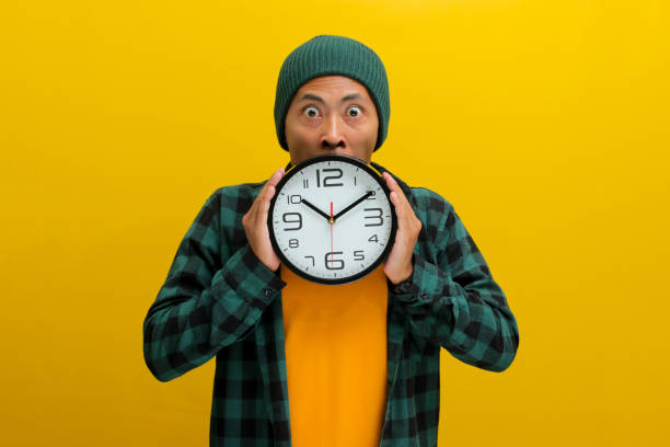 istock an Asian man holds a clock in front of his face, looking at the camera with a surprised expression 2152143519