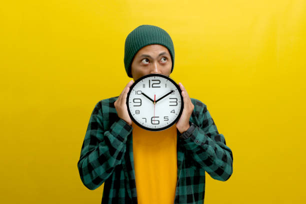 istock Pensive Asian man, looking aside at an empty copy space while holding a clock in front of his face 2152156018