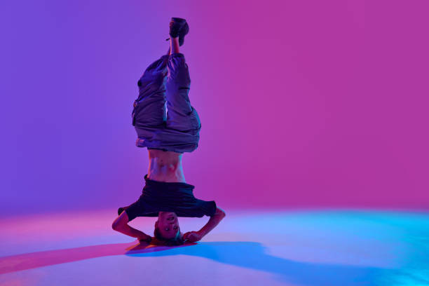 Stylish dressed young dancer, guy performing spinning on head in mixed neon light against vibrant gradient background. Concept of sport and hobby, music, fashion and art, movement. Ad