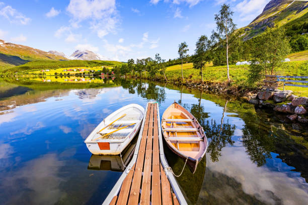 Wooden pier with boats. Shallow lake Eidsvatnet in the mountains of Norway. The water reflects lush clouds, blue skies and forested shores. Summer in Scandinavia.