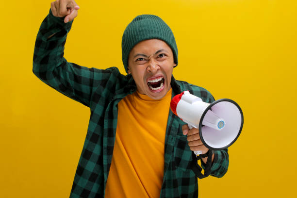 istock Angry Asian Man Yells into Megaphone, Demanding Change or Protesting, Isolated on Yellow Background 2152175891