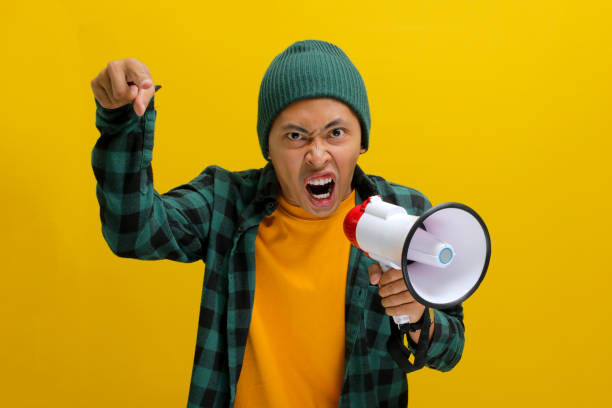 istock Angry Asian Man Yells into Megaphone, Demanding Change or Protesting, Isolated on Yellow Background 2152176024