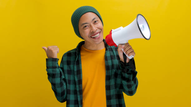 istock Happy Asian Man in Beanie Talks into Megaphone, Points to Copy Space for Discount Promotion or Sale 2152181212