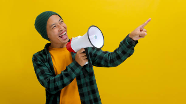 istock Happy Asian Man in Beanie Talks into Megaphone, Points to Copy Space for Discount Promotion or Sale 2152181224