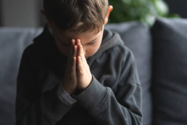 istock Young Boy Sitting on a Sofa With Hands Clasped in a Moment of praying 2152185564