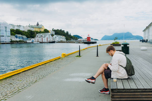 Side view of a male tourist with backpack sitting by the sea in Alesund city surfing Internet by his phone during summer day