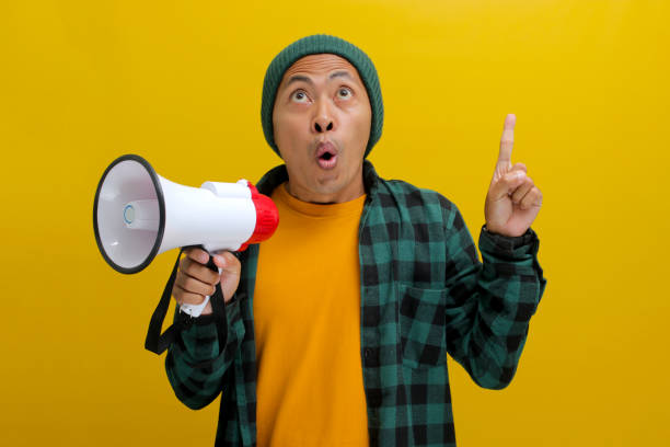 istock Happy Asian Man in Beanie Talks into Megaphone, Points to Copy Space for Discount Promotion or Sale 2152191059