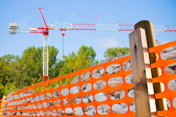 Tower crane in a construction site with metal scaffolding to work on the building facade and perforated safety orange plastic grid to delimit and protect construction site areas.