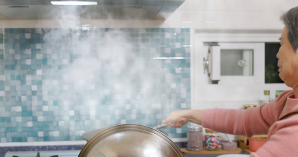 asian elderly woman is blowing the lid off from wok when cook with much oil smoke in kitchen - one of causes for lung cancer
