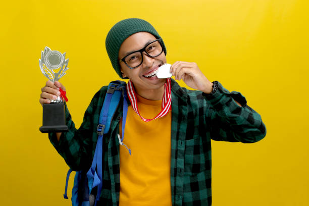 istock Young Asian Student with Backpack Bites Medal and Holds Silver Trophy on Yellow Background 2152217479