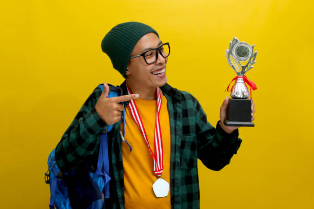 istock Young Asian Student Points at His Winning Trophy While Carrying a Backpack on Yellow Background 2152218121