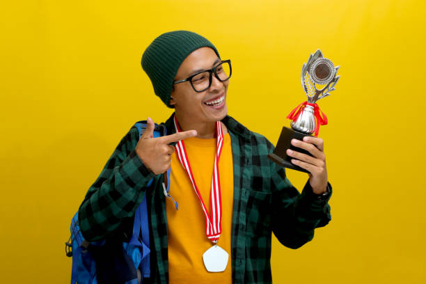 istock Young Asian Student Points at His Winning Trophy While Carrying a Backpack on Yellow Background 2152218190