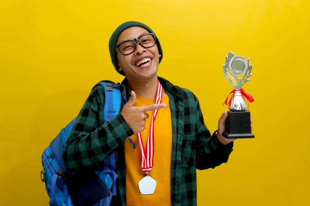 istock Young Asian Student Points at His Winning Trophy While Carrying a Backpack on Yellow Background 2152218772