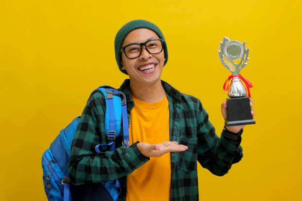 istock Asian Student Celebrates Success with Trophy on Yellow Background 2152222814
