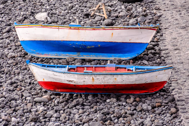 Two boats are sitting on a rocky beach. One is blue and the other is red. The boats are old and rusted