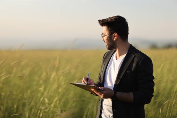 A young agronomist holds a folder in his hands on a green wheat field. A farmer makes notes on the background of agricultural land during sunset.