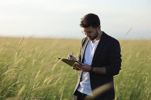 A young agronomist holds a folder in his hands on a green wheat field. A farmer makes notes on the background of agricultural land during sunset.