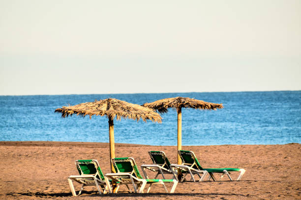 Two umbrellas and four lounge chairs are on a beach. The beach is empty and the sky is blue