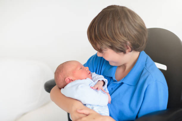 Happy laughing boy holding his sleeping newborn baby brother. Siblings with big age difference. Children playing at home on a white couch. New born child taking a nap. Kids bonding. Family love.