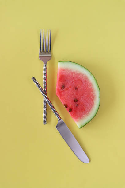 Stock photo showcases a creative watermelon clock, symbolizing the concept of intermittent fasting and healthy eating. Surrounding the clock are various nutritious foods, accompanied by a knife and fork, emphasizing balanced nutrition and mindful eating habits essential for overall well-being.