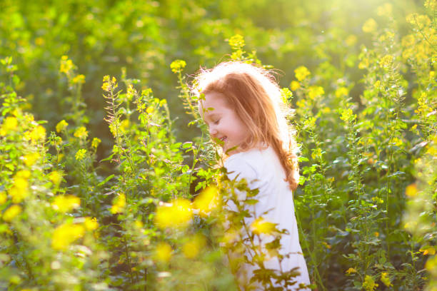 Cute little girl in white dress playing in blooming spring park with first white wild anemone flowers. Child on Easter egg hunt in blooming garden. Kids play outdoor picking flower bouquet.