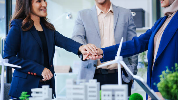 istock Three businesspeople gather at a desk, symbolizing unity with joined hands. Led by a middle-aged Asian man and woman, they strategize sustainable solutions for a cleaner, energy-efficient future. 2152294843