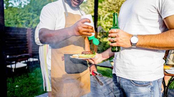 Young people preparing a barbecue in the backyard and having a great time, a man with an apron grilling and giving burgers to his friends