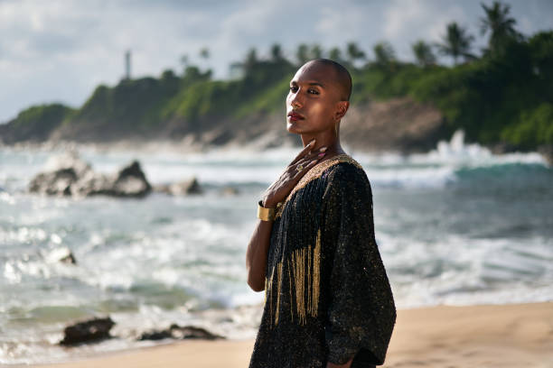 Outrageous lgbtq black model in luxury gown poses on scenic ocean beach with moody fashion look. Non-binary ethnic model in long posh dress and accessories looks at camera arrogantly, touches a neck. escandalosa modelo negra lgbtq con vestido de lujo posa en la pintoresca playa del océano con un aspecto de moda malhumorado. modelo étnica no binaria con vestido largo y elegante y accesorios mira a la cámara con arrogancia, toca el cuello.
