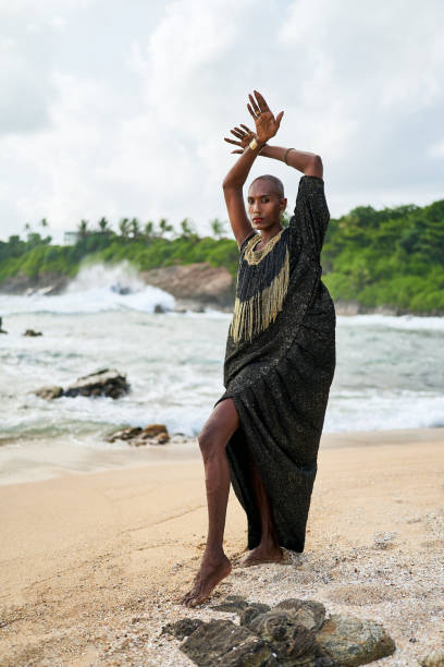 istock Epatage lgbtq black male posing with hands up on camera on scenic ocean beach. Non-binary ethnic fashion model in long posh dress wears jewellery stands gracefully on sea shore and lighthouse. 2152307360
