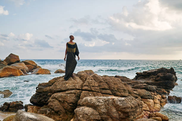 istock Non-binary black godlike person poses gracefully standing on rocks in ocean. Trans ethnic fashion model in posh dress and jewellery on rocky beach by storm . Lgbtq. Pride month 2152307553