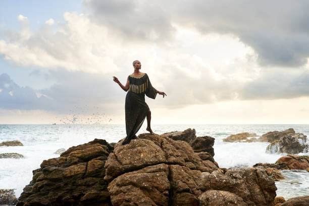 Non-binary black godlike person poses gracefully standing on rocks in ocean. Trans ethnic fashion model in a posh dress and jewellery on rocky beach by storm . Lgbtq. Pride month