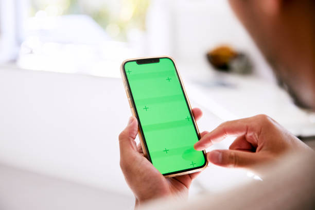 Rear view of a man holding his green screen phone In his bright kitchen, a man stands, his back to the camera, holding up his green-screen phone man looking at phone over shoulder stock pictures, royalty-free photos & images