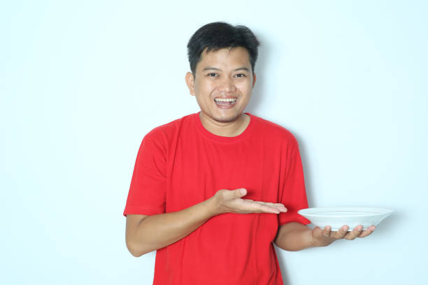 istock Young Asian man smiling happy while showing empty dinner plate with his open palm. Wearing red a shirt 2152317185