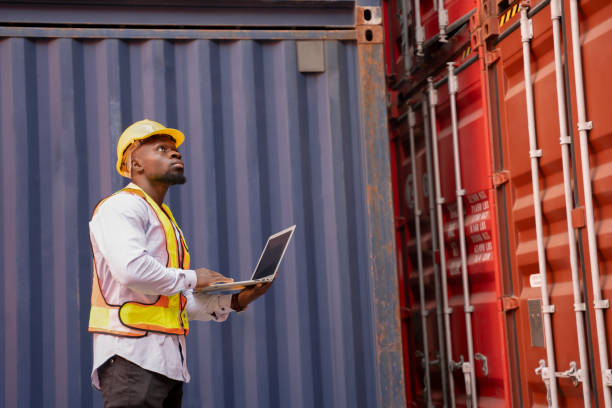 istock black african man wears hardhat and vest holding laptop computer checking electronic seal on container cargo,young male technician working in shipping yard 2152329320