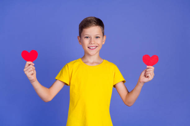Photo of cheerful adorable cute small boy wear stylish yellow clothes hold red paper postcards isolated on purple color background Photo of cheerful adorable cute small boy wear stylish yellow clothes hold red paper postcards isolated on purple color background. cute boy show love symbol hand stock pictures, royalty-free photos & images