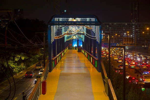 Illuminated footbridge at Bangkok Linear Park parallel to Duang Phithak Rd at night. On road is traffic jam going south. Bridge and footbridge is decorated with lights.