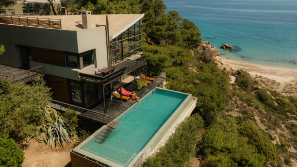 Photo of a luxury villa on a cliff that overlooks the sea. A young woman is sunbathing on the edge of the infinity pool
