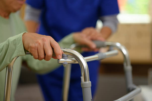 Caregiver or physiotherapist supporting elderly patient using walking frame at nursing home.