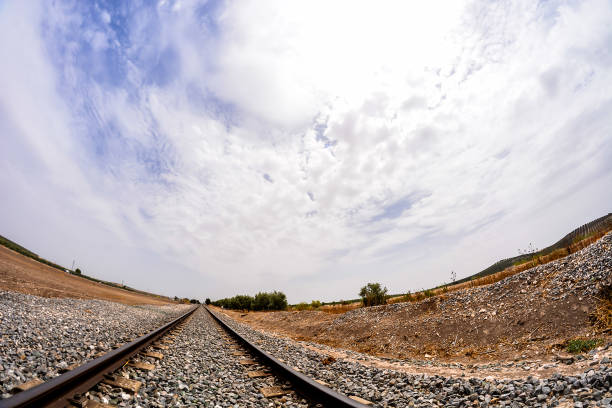 The sky is cloudy and the sun is shining through the clouds. The train tracks are in the foreground and the rest of the image is a wide shot of the countryside