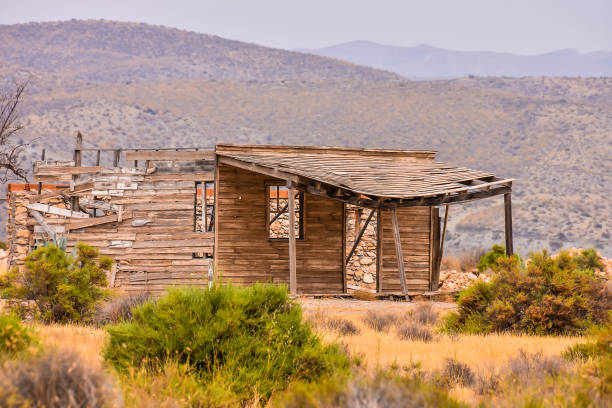 A dilapidated old house sits in a barren desert landscape. The house is in a state of disrepair, with broken windows and a roof that is missing. The surrounding area is dry and barren