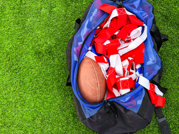 A large athletic duffel bag is filled with equipment for flag football play. Red and blue flags and a football fill the bag. Green turf football field background.