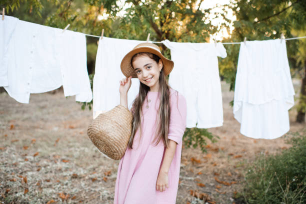 Smiling child girl 6-7 year old wearing straw hat and linen dress holding basket standing over clothesline with dry clothes outdoor. Looking at camera. Summer season.