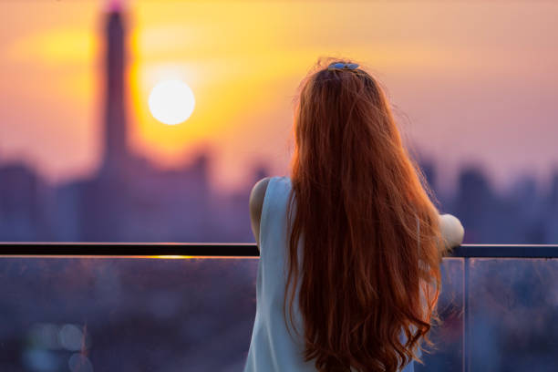 istock Woman looking and enjoying sunset view from balcony with the sun setting behind skyscraper in busy urban downtown with loneliness for solitude, loneliness and dreaming of freedom lifestyle 2152513748