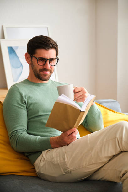 istock A man is sitting on a couch reading a book and drinking coffee. 2152567776