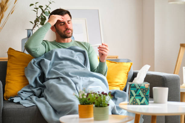 istock A man with fever is sitting on a couch with a tissue box and a thermometer. 2152568533