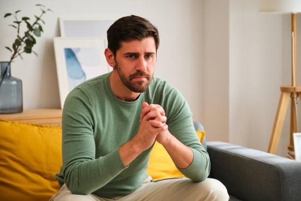 istock Sad man is sitting on a couch with his hands clasped in front of him. 2152569037