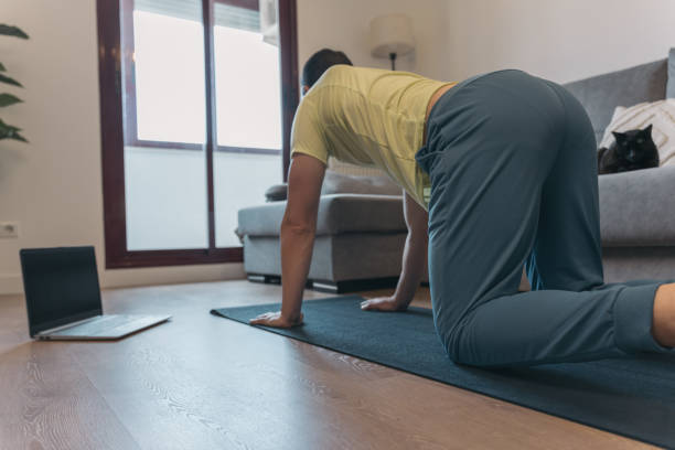 istock Anonymous Man Stretching on Mat with Laptop for Online Class in Home Setting. Unrecognizable man does yoga stretches on mat with laptop nearby for online class in cozy home environment. 2152577108