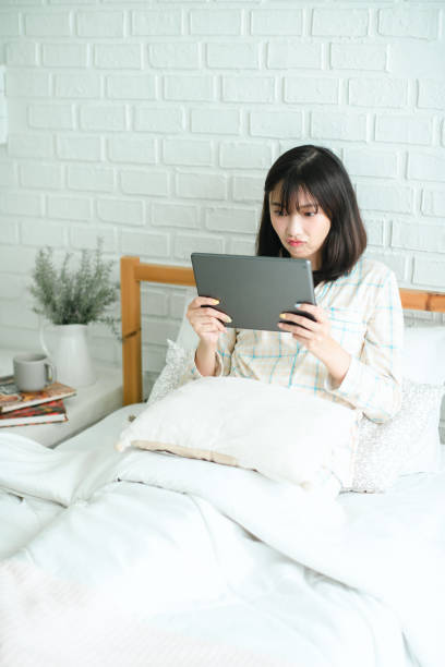 istock Thoughtful young female in pajamas browsing tablet while lying on bed in her bedroom 2152584053