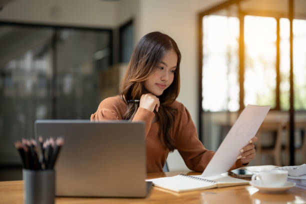 istock A young professional woman smiles while examining papers in a well-lit, modern office setting, with a laptop. 2152609669